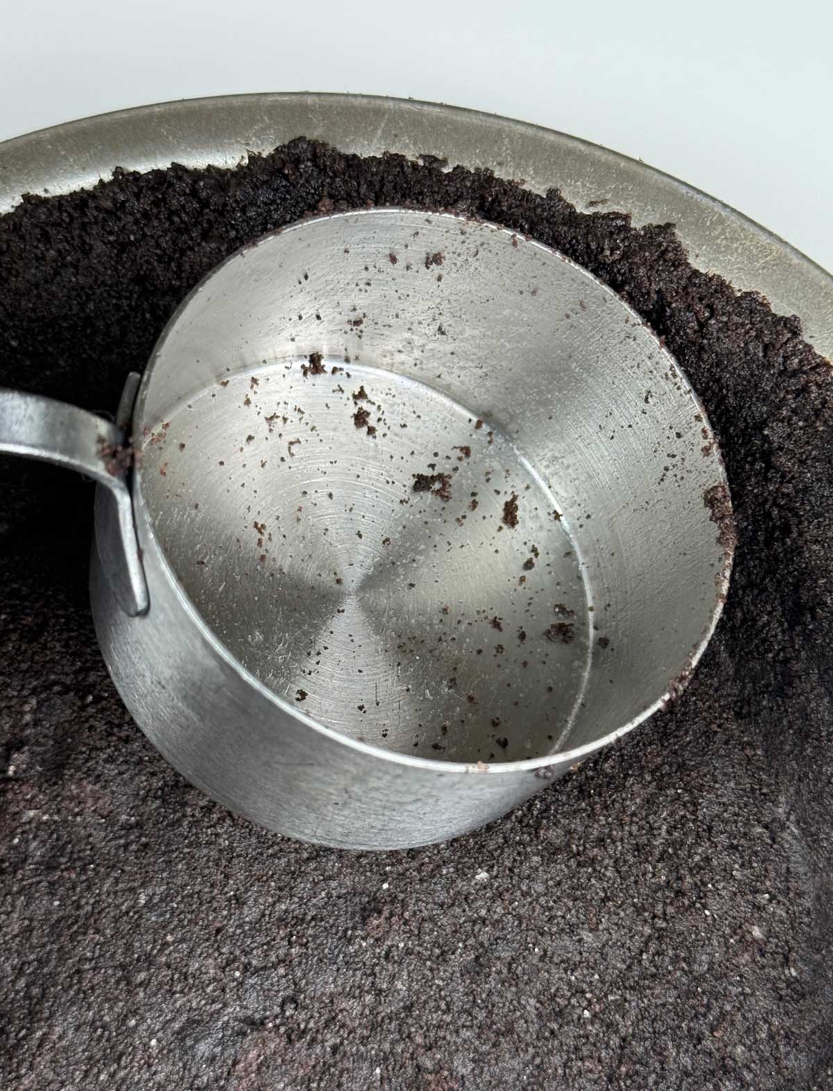 Oreo crumbs in a pie dish being pressed into a 9-inch pie plate with a measuring cup.